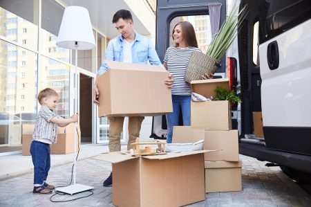 Portrait of happy young family holding cardboard boxes standing next to moving van and smiling looking at their son